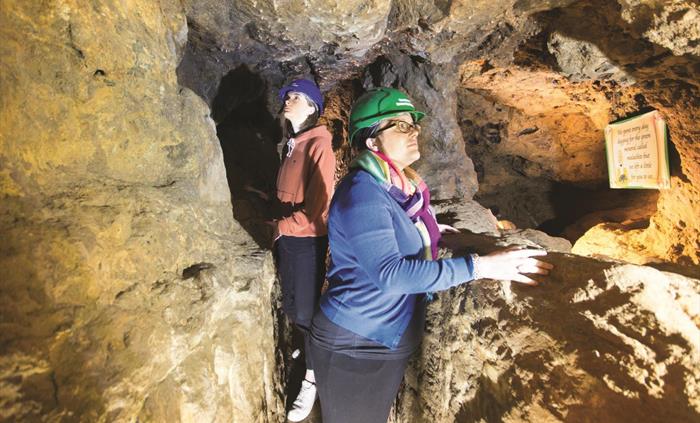 Two people standing in the underground tunnel, facing away from each other, looking at the rock walls of the mine.